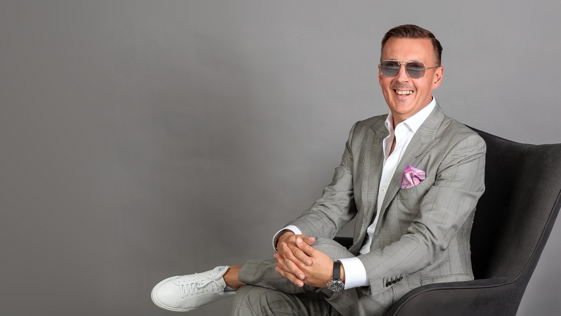 Ing. Christian Weinberger-Prammer im Querformat, lächelnd in einem dunklen Sessel sitzend, in hellgrauem Karo-Anzug mit rosa Einstecktuch und Sonnenbrille, vor grauem Studiohintergrund.